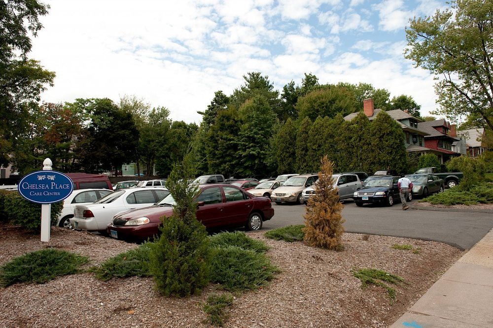 A parking lot filled with cars surrounded by trees and greenery under a partially cloudy sky.