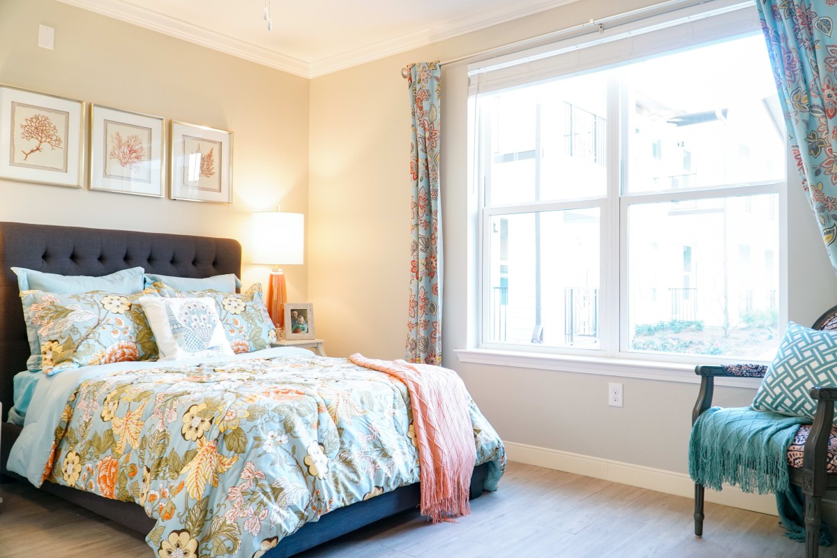 A well-lit bedroom featuring a comfortable bed with floral bedding, a bedside lamp, and large window.