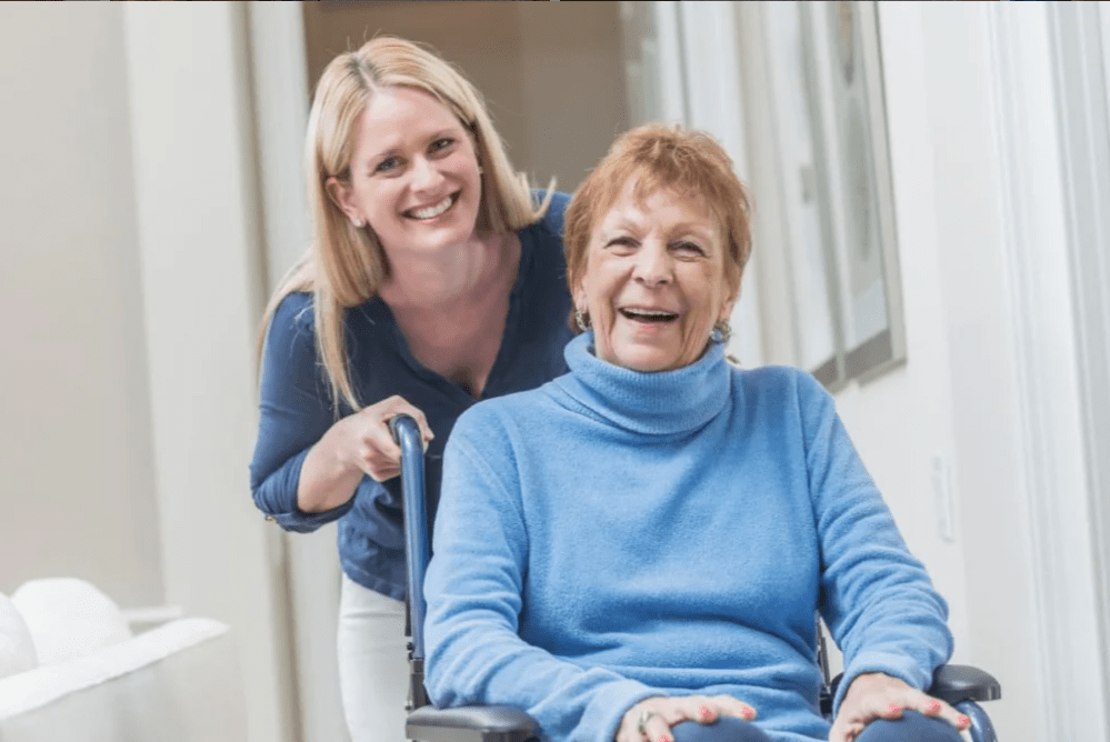 A caregiver with an elderly woman in a wheelchair, both smiling indoors.