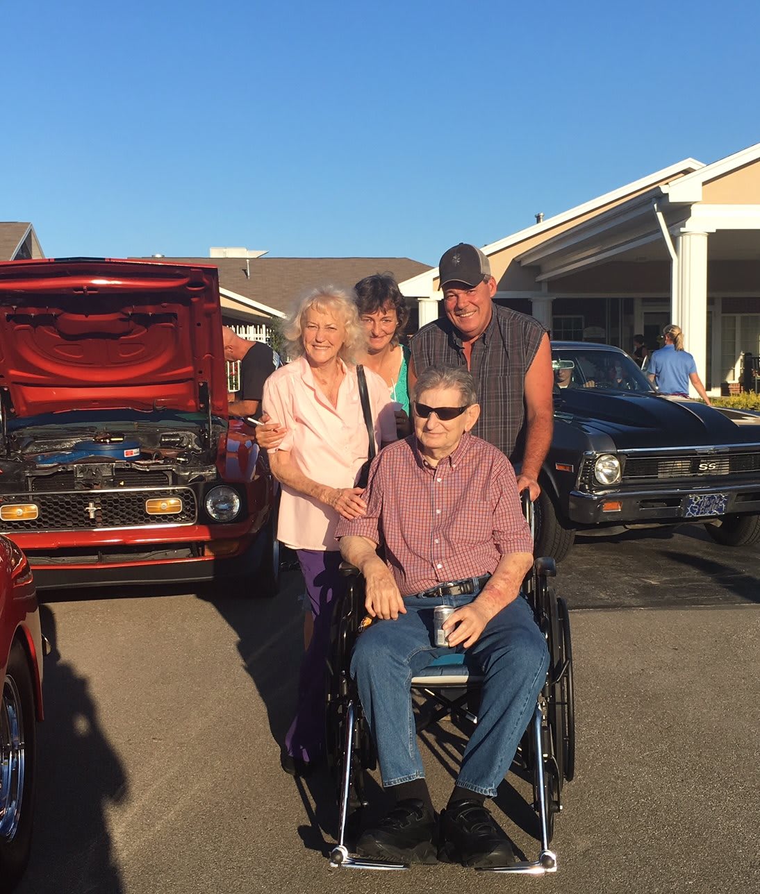A group of people posing outdoors in front of cars, with one person in a wheelchair.