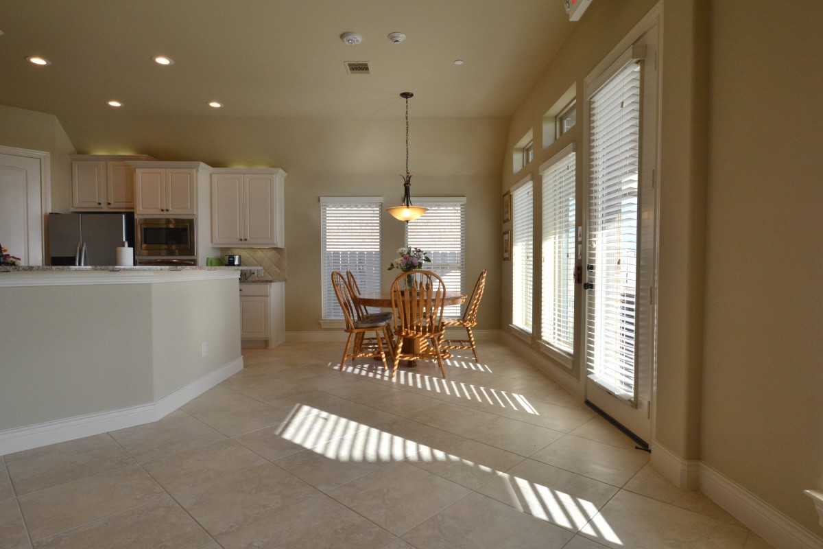 A bright kitchen and dining area with natural light streaming through blinds, featuring a wooden table and chairs on tiled flooring.