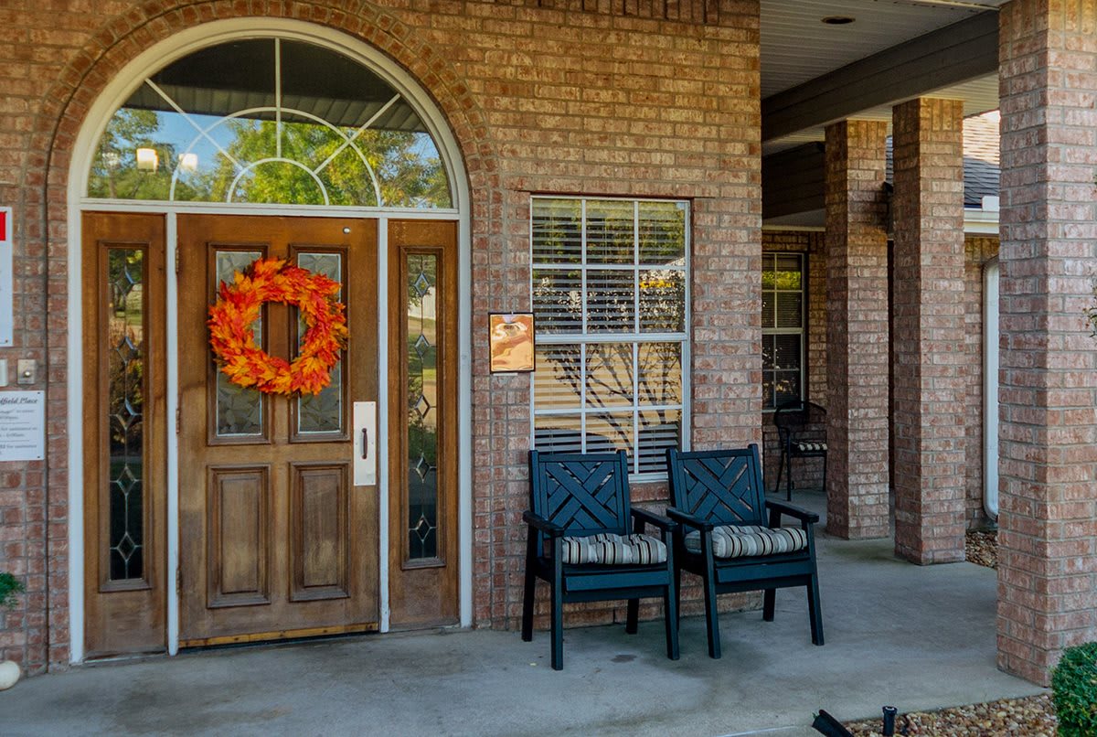 A cozy porch with a wooden door adorned with an orange wreath, flanked by two chairs against a brick wall.