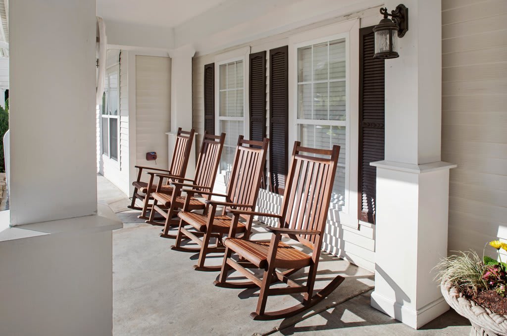 A sunny porch featuring several wooden rocking chairs lined up in front of large windows.