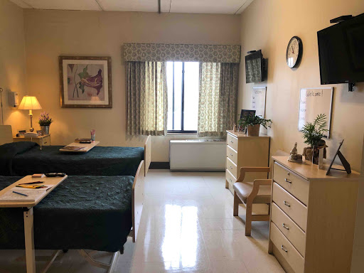 A well-lit hospital room featuring two patient beds, a window with curtains, and a simple dresser with decorative items.