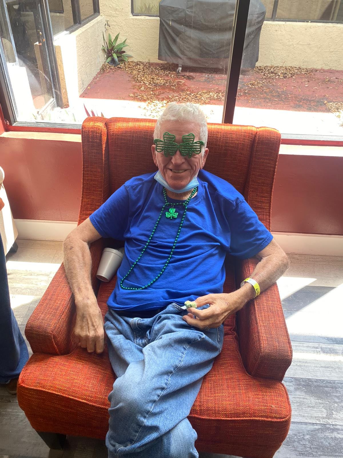 An elderly man wearing sunglasses and a blue shirt is seated comfortably in an armchair indoors.