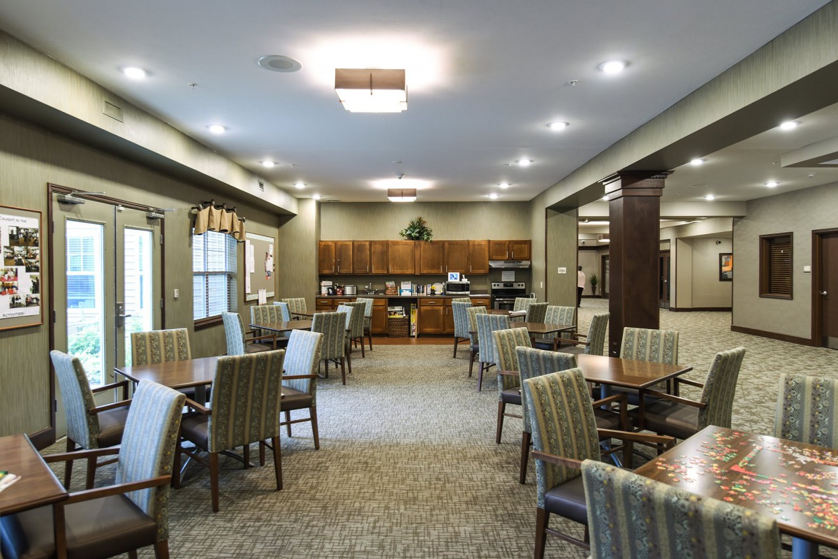 A spacious dining area with multiple tables and chairs, featuring warm lighting and an open kitchen setup.