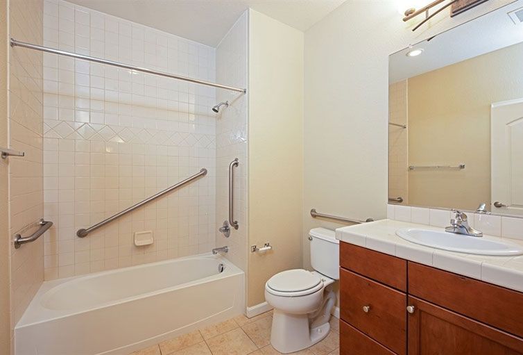 A clean and modern bathroom featuring a bathtub with grab bars, a toilet, and a sink with a mirror.