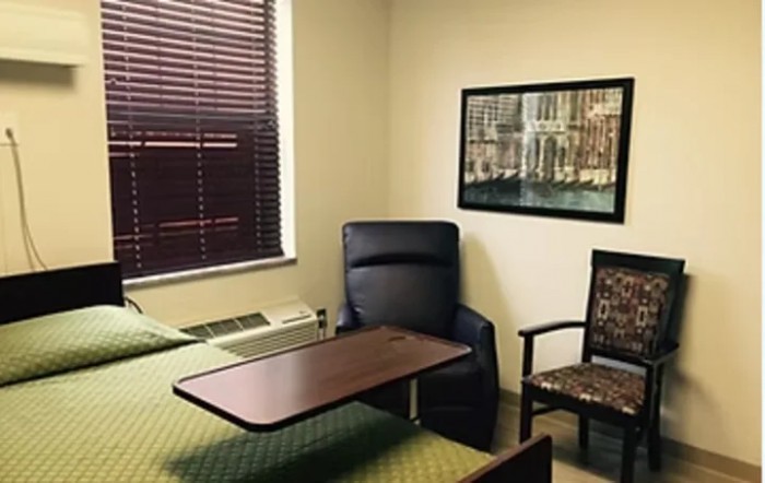 A small room with a bed, a reclining chair, and a table near the window.