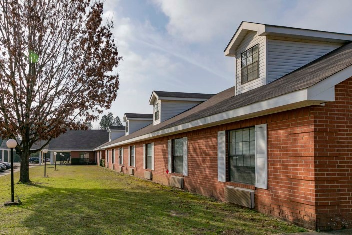 A long brick building with multiple windows and a sloped roof sits beside a green lawn and tree on a clear day.