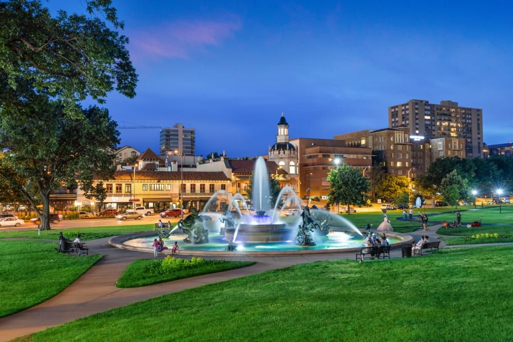 A beautifully lit fountain in a city park at dusk, with buildings and lights in the background.
