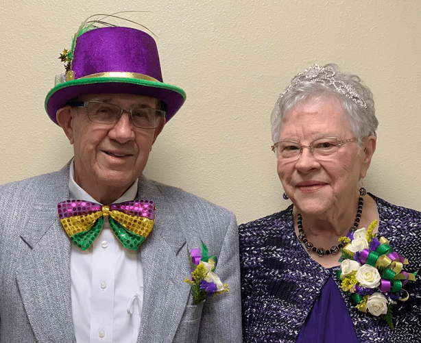 An elderly couple dressed in formal attire, with the man wearing a colorful hat and bow tie, and both adorned with floral accessories.