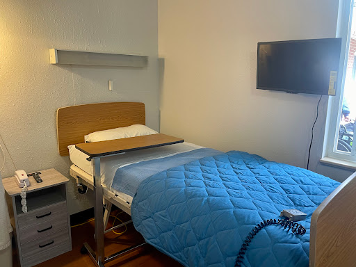 A clean and minimalistic hospital patient room featuring a bed with a blue blanket and a television.