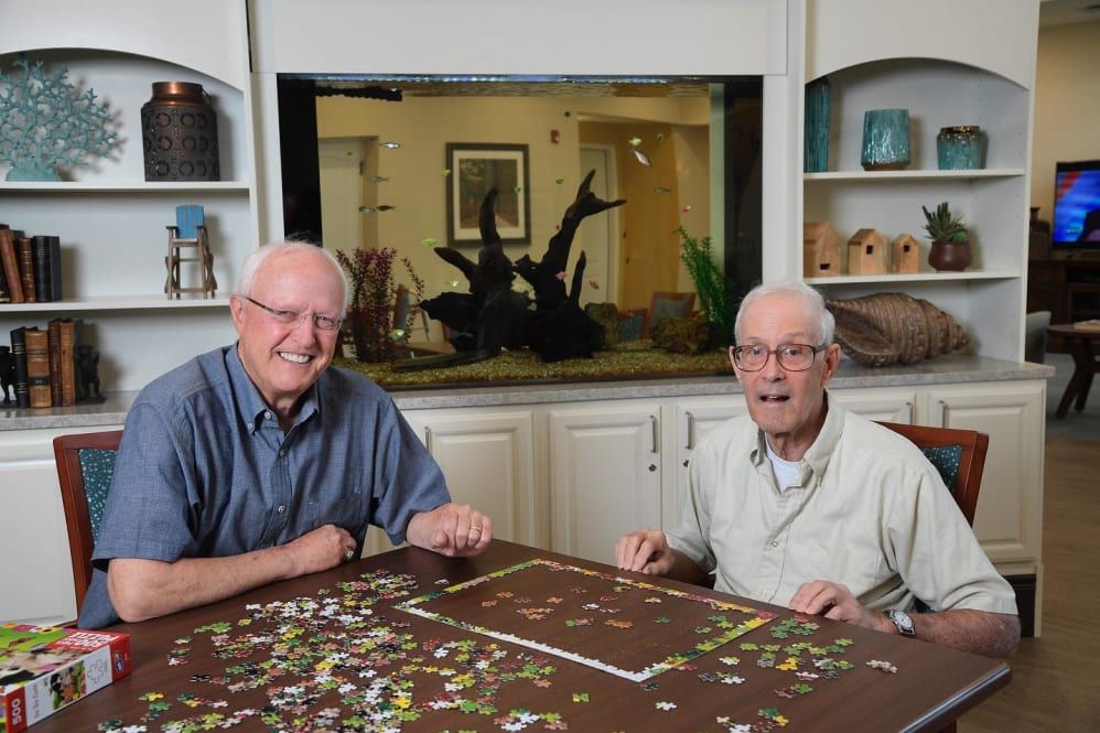 Two elderly men working on a puzzle near an aquarium.