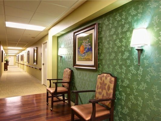 A hallway with chairs and artwork on green wallpapered walls, illuminated by wall lamps.