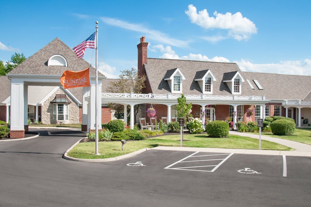 A large house with a driveway and flags is surrounded by well-maintained landscaping on a sunny day.