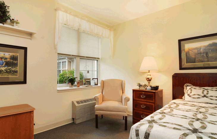 A cozy bedroom with a comfortable chair by the window and a lamp on the dresser.