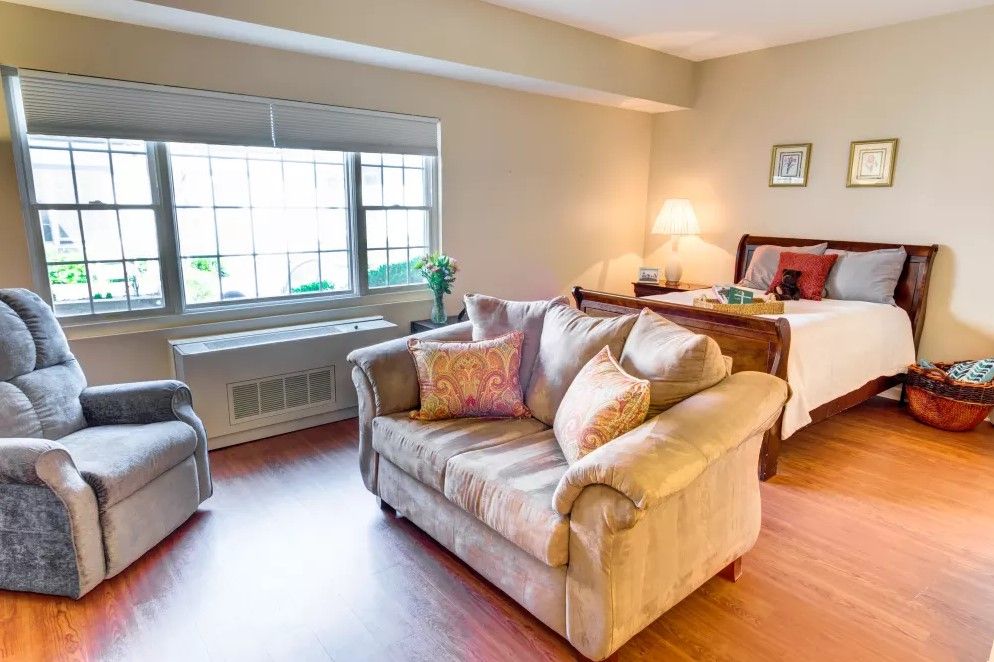 A cozy living space with a sofa, armchair, and bed, featuring warm lighting and hardwood floors.