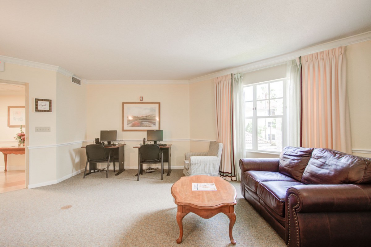 A spacious living room with a large sofa, coffee table, desks, and chairs, featuring a large window allowing natural light to fill the space.