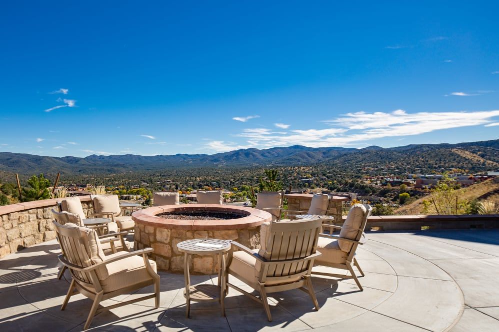 A scenic outdoor patio with wooden chairs surrounding a fire pit, overlooking a mountainous landscape under a clear blue sky.