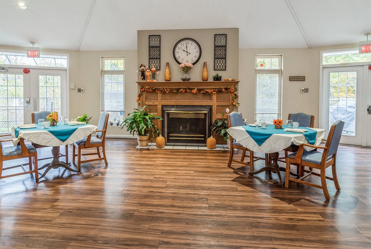 A cozy dining room with wooden floors, tables set for meals, a central fireplace, and a clock above the mantel.