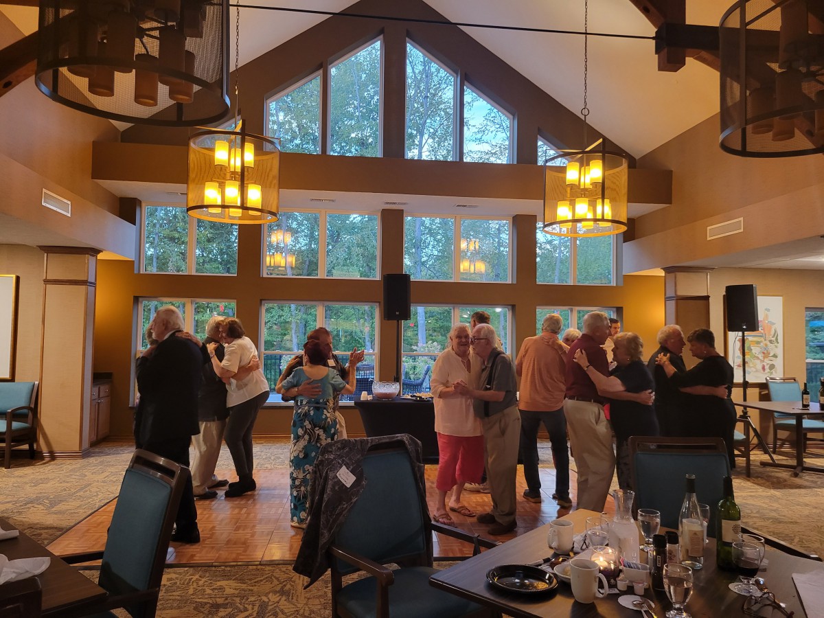A group of people dancing at a social gathering in a beautifully lit room with large windows.