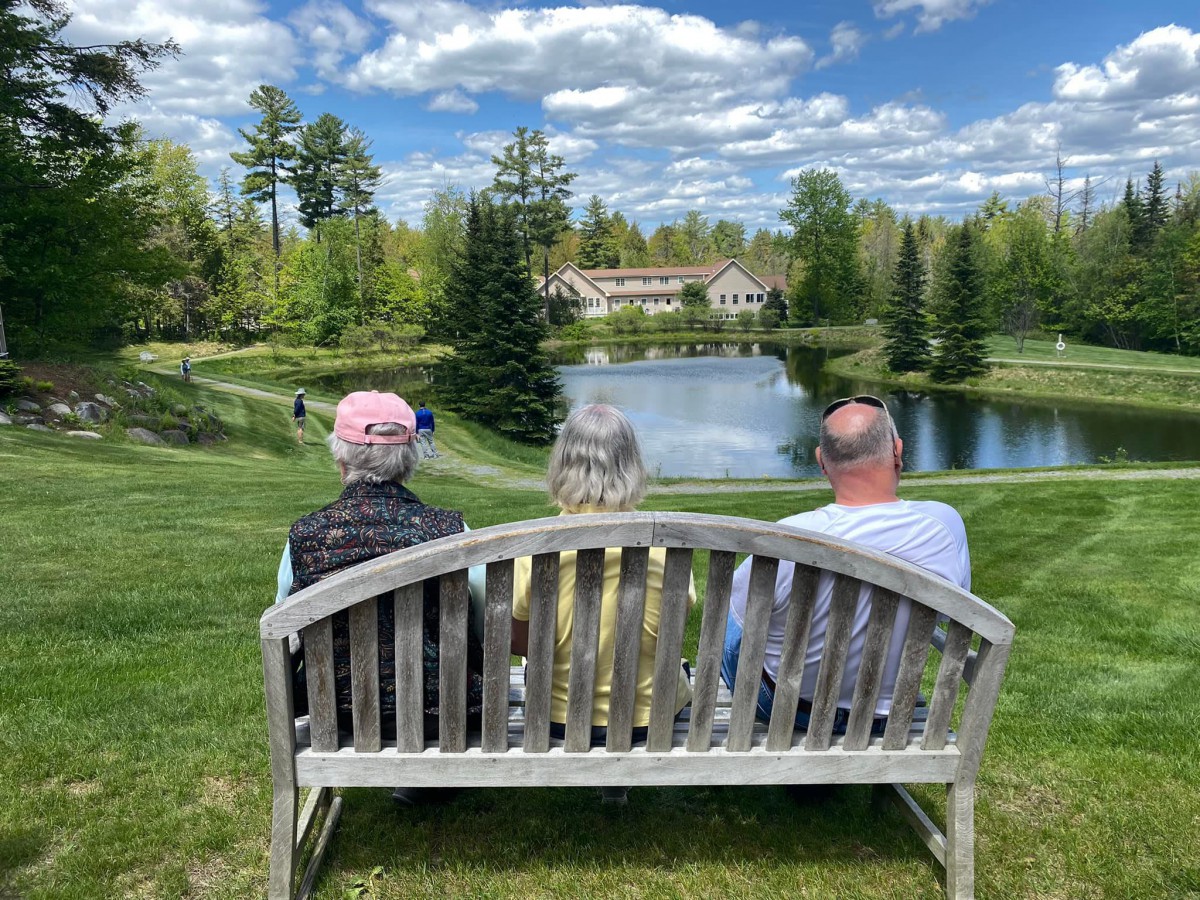 Three people sitting on a bench enjoying a scenic view of a pond and lush landscape.