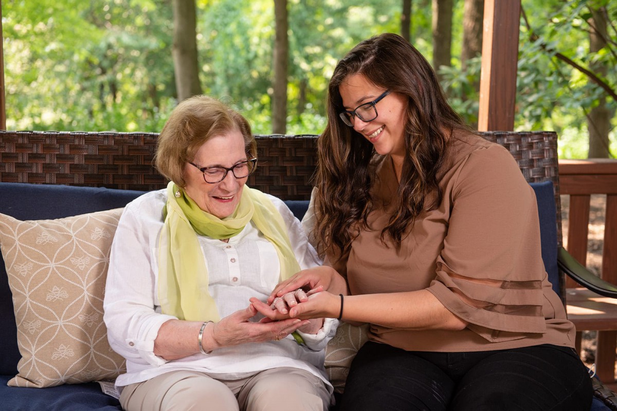 A caregiver assists an elderly woman indoors, both smiling warmly.