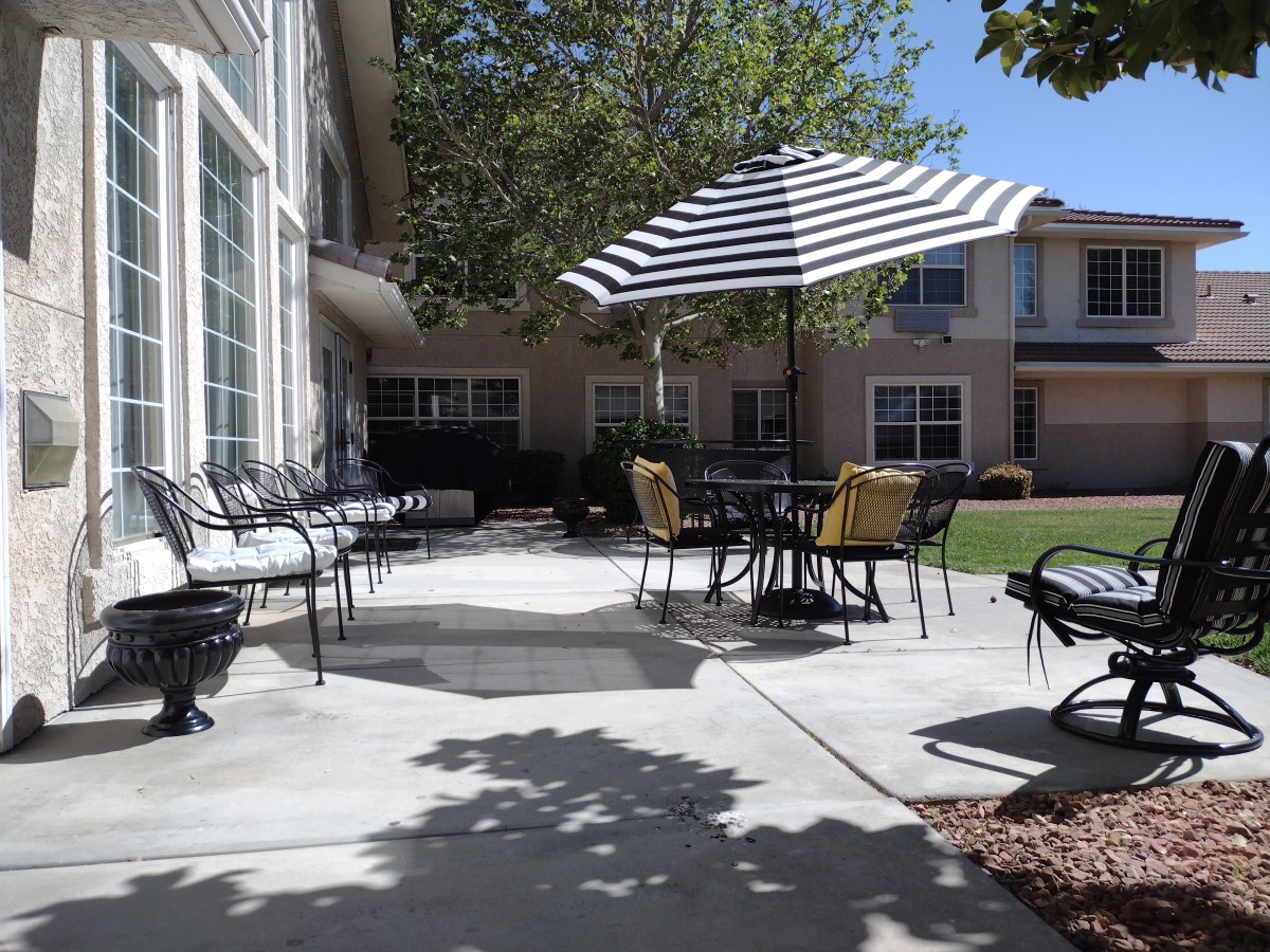 A sunny patio area with tables, chairs, and a striped umbrella surrounded by a garden and buildings.