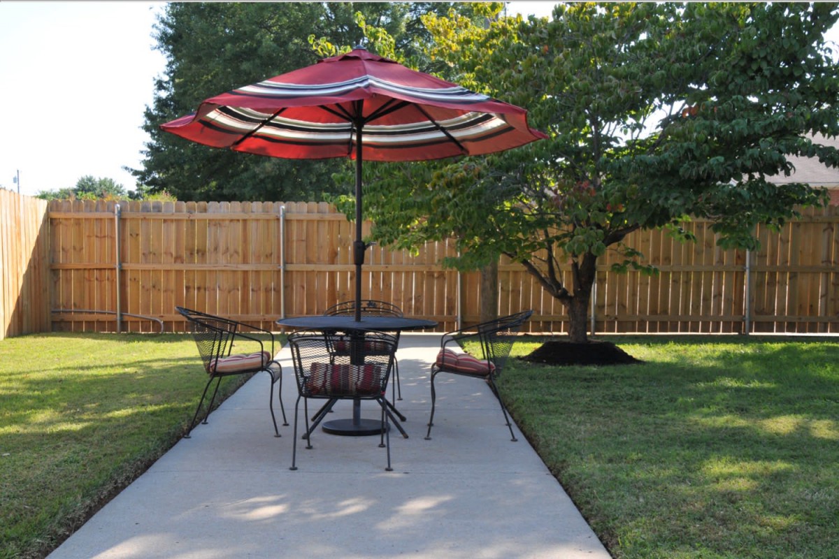 A patio with a table and chairs under a red umbrella, surrounded by a lush garden.