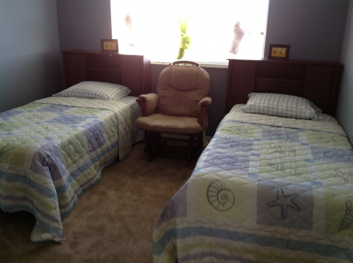 A cozy bedroom features two twin beds with patterned quilts and a comfortable chair between them, illuminated by light from the window.