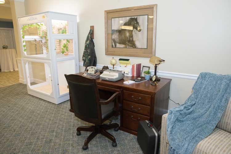 An office setup featuring a wooden desk with a typewriter, telephone, and lamp, accompanied by an office chair. A framed artwork hangs above the desk.