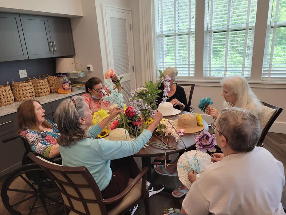 A group of elderly women enjoying a gathering around a table with floral decorations, wearing festive hats indoors.