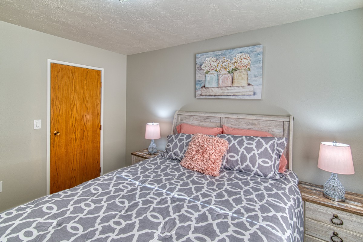 A cozy bedroom with a neatly made bed featuring patterned bedding, a bedside lamp, and artwork on the wall.