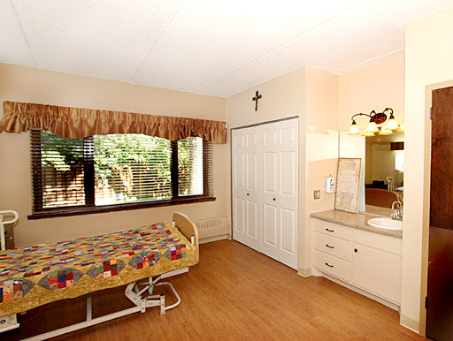 A well-lit bedroom featuring a hospital-style bed, large windows, and a simple wooden floor.