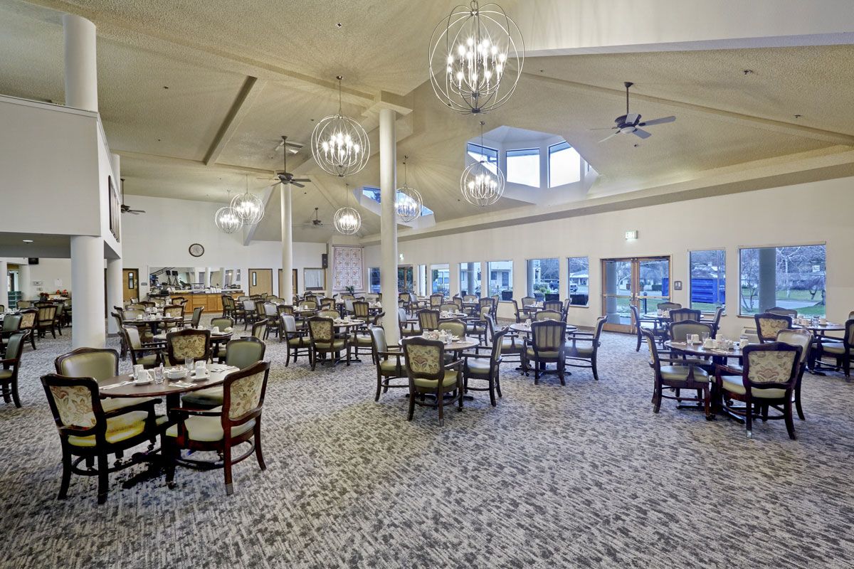 A spacious dining room with elegantly arranged tables and chairs, featuring chandeliers and large windows.