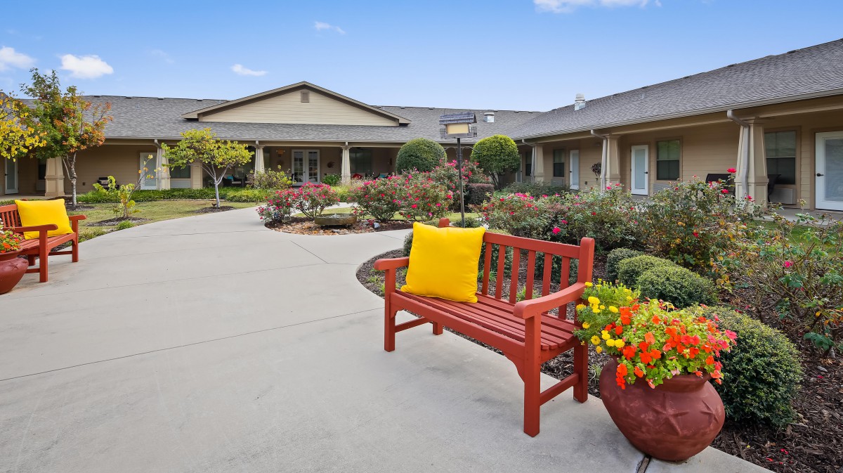A tranquil outdoor area with a red bench and yellow cushion, surrounded by flowering plants and a well-maintained garden in front of a building.