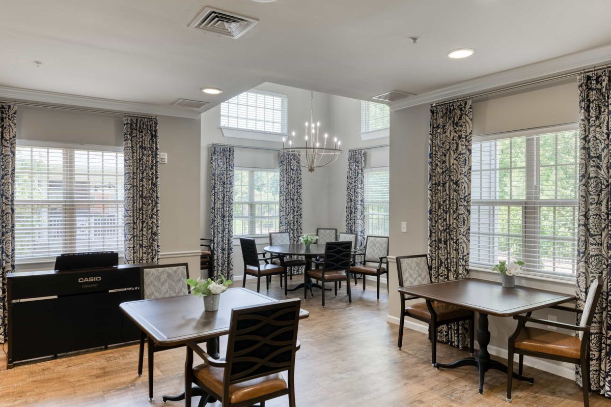 A well-lit dining room with multiple tables and chairs, large windows adorned with curtains, and a chandelier hanging from the ceiling.