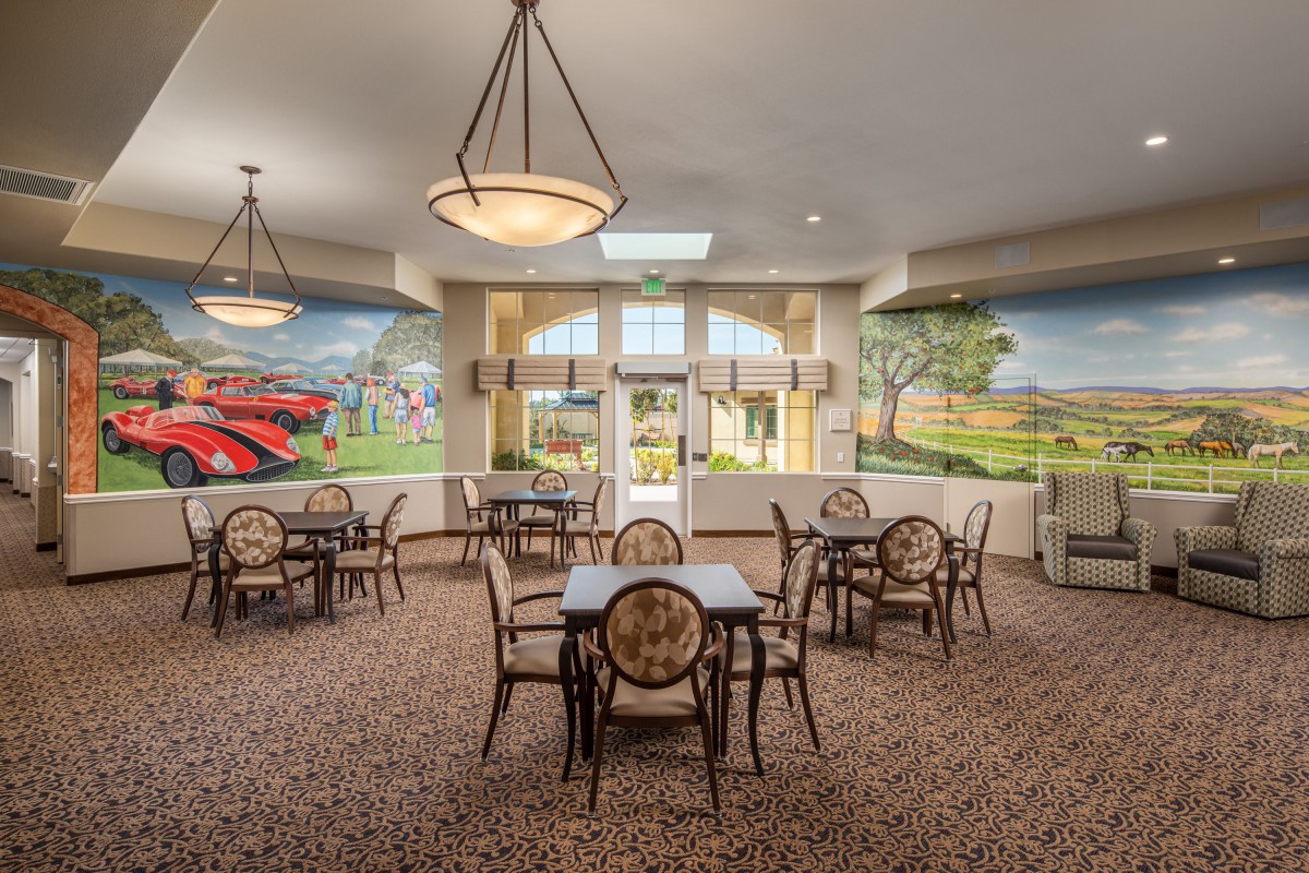 A spacious dining area featuring several tables and chairs, illuminated by chandeliers, with colorful murals on the walls.