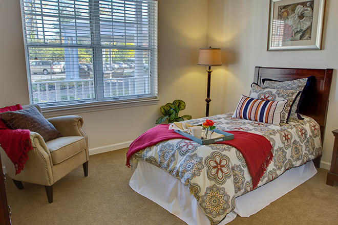 A cozy bedroom with patterned bedding, a floor lamp, and an upholstered chair near the window.