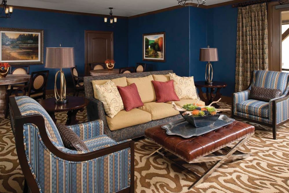 A stylish living room with blue walls, featuring a comfortable sofa and armchair, accented by red and beige cushions. A patterned rug complements the furniture, while lamps and artwork add decorative touches.