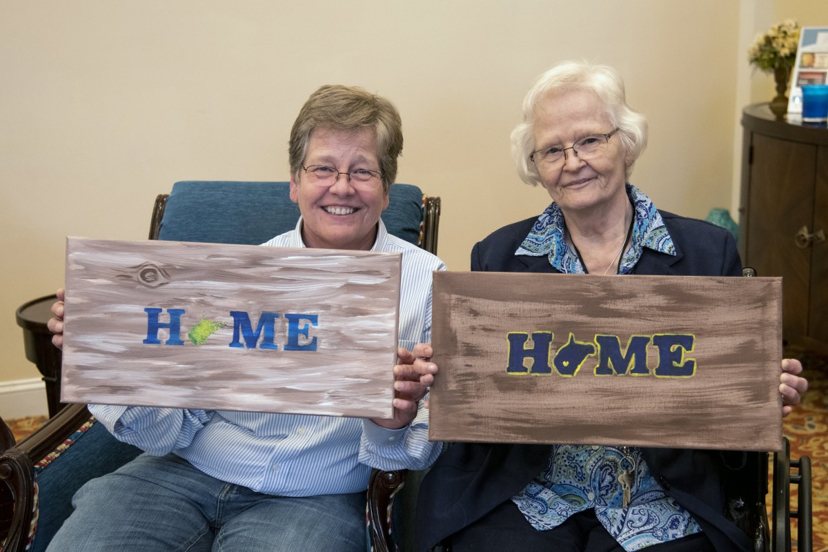 Two elderly women smiling and holding 'HOME' signs indoors.