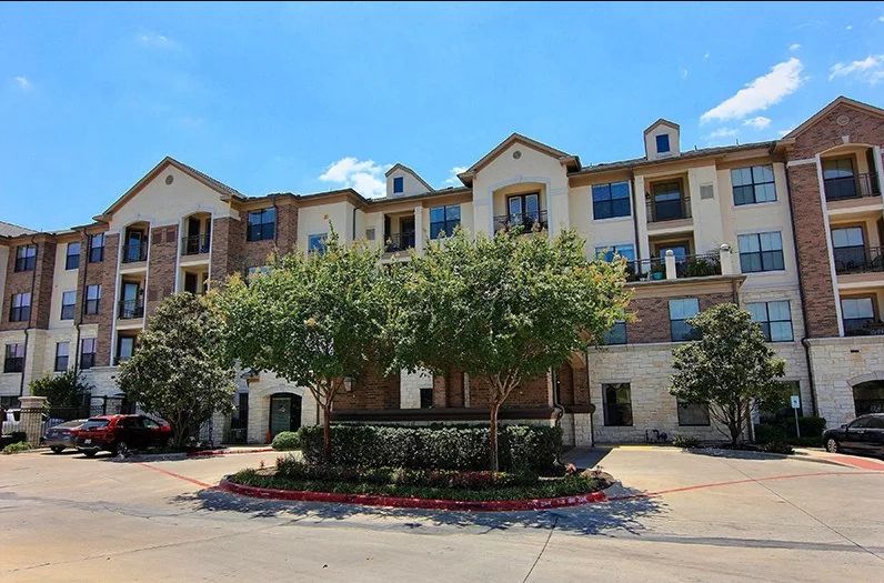A multi-story residential apartment building surrounded by trees under a clear blue sky.