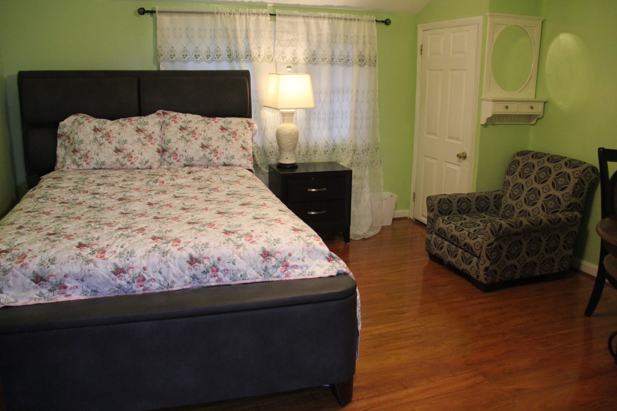 A cozy bedroom with a bed, nightstand, and armchair, featuring a lamp and curtains on the window.