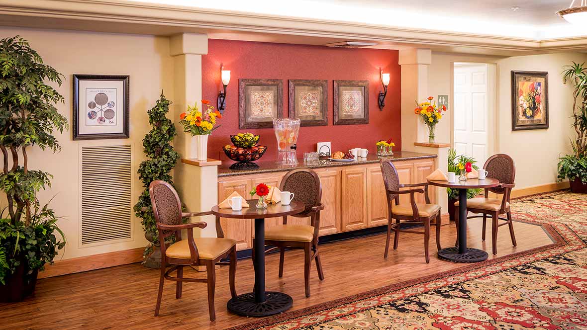 A cozy cafe interior with tables and chairs, decorated with flowers and paintings, featuring a beverage station.