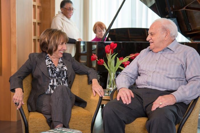 Two people are engaged in a conversation while sitting in a room with a piano and flowers.