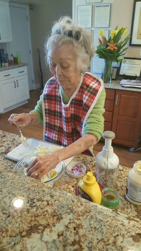 An elderly woman preparing food in the kitchen, wearing an apron and surrounded by cooking ingredients.
