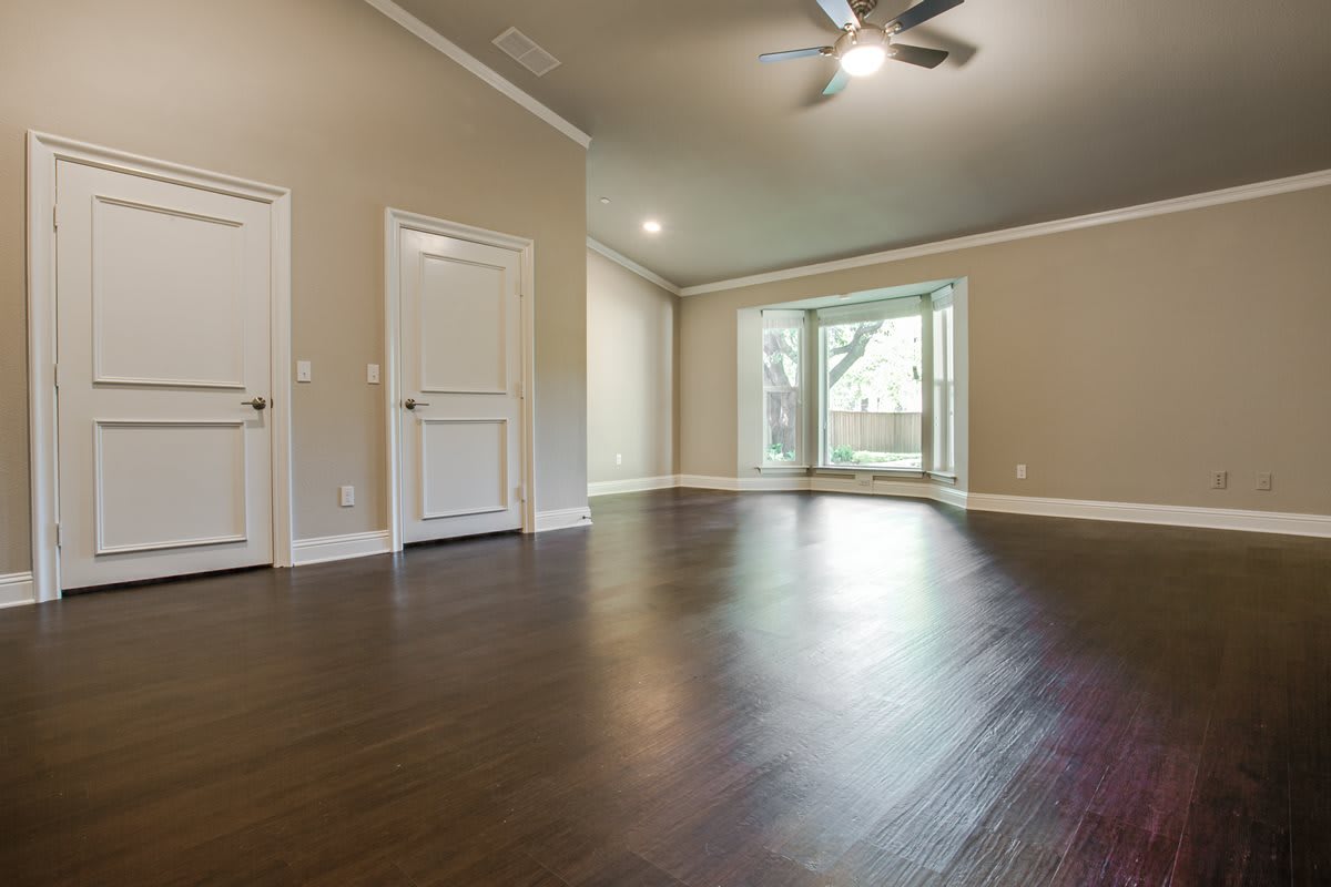 A spacious, empty room with hardwood flooring and a ceiling fan, illuminated by natural light from large windows.