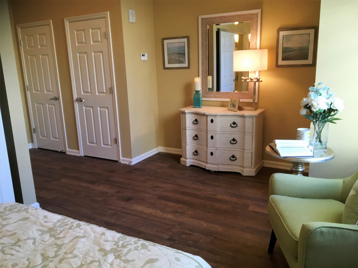A cozy bedroom with wooden flooring, featuring a dresser, lamp, and artwork on the wall.