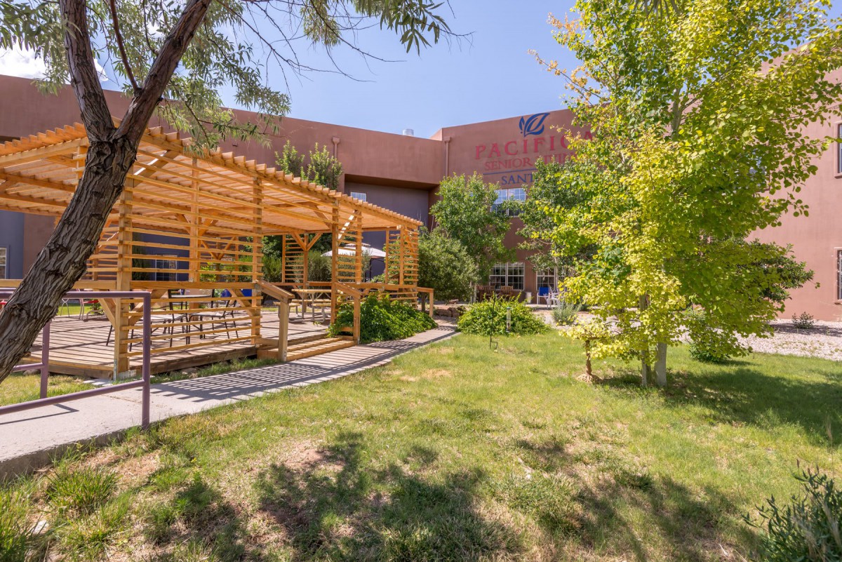A sunny outdoor space featuring a pergola, surrounded by lush greenery and trees, with a building in the background.