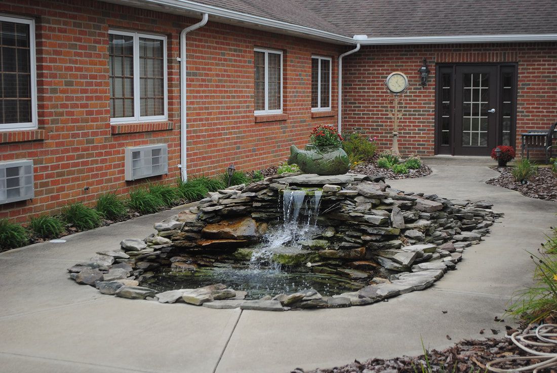 A serene stone-bordered pond with a cascading waterfall located in a courtyard next to a brick building.